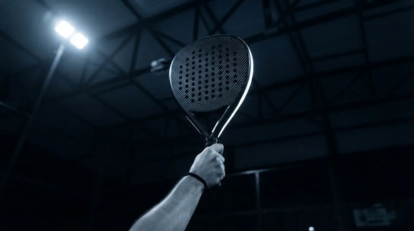 A diamond-shaped padel racket raised at the apex of a smash under floodlights