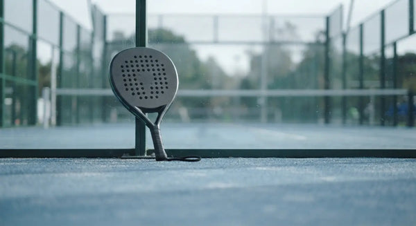 A round padel racket resting against the glass wall of an empty padel court